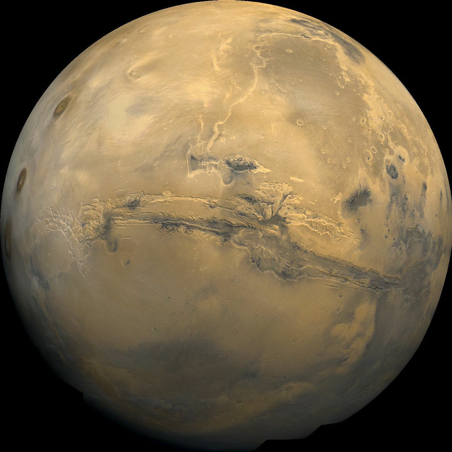 Der Mars ist bildfüllend und detailreich abgebildet. Quer über die Mitte der Vorderseite verläuft eine tiefe Schlucht, parallel dazu einige Rillen. Links und rechts sind runde, dunkle Flecken zu sehen, es sind Vulkangipfel.