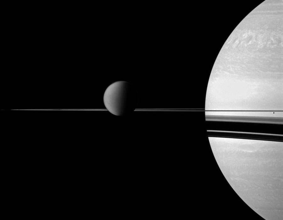 Rechts ragt Saturn ins Bild, in der Bildmitte verlaufen waagrecht die Ringe. Links neben Saturn ragt Titan über die Ringe, rechts ist der winzige Mond Enceladus.