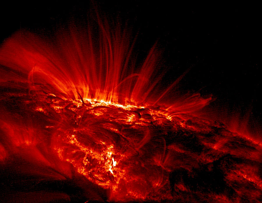 Vor einem dunklen Hintergrund glühen einige Stellen auf der Sonne wie Lava, von diesen steigen rote Schleifen entlang von Magnetfeldlinien auf.