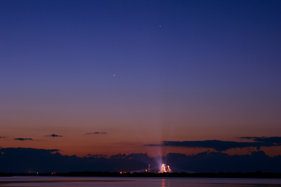 Unter einem violettblauen Himmel mit einem dunkelroten Streifen über dem Horizont steht vor einer dunklen Wolkenbank die hell erleuchtete Startrampe mit der startbereiten Raumfähre. Nach oben leuchten Scheinwerfer, die auf die Raumfähre gerichtet sind.