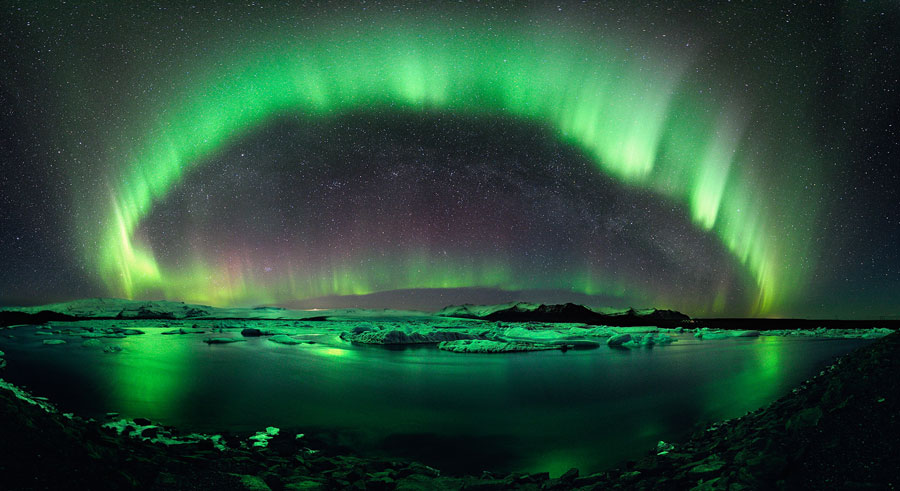 Über einem ruhigen See und einem Gebirge dahinter breitet sich ein hellgrün leuchtender Polarlichtbogen aus. Die grünen Lichter spiegeln sich im Gewässer.