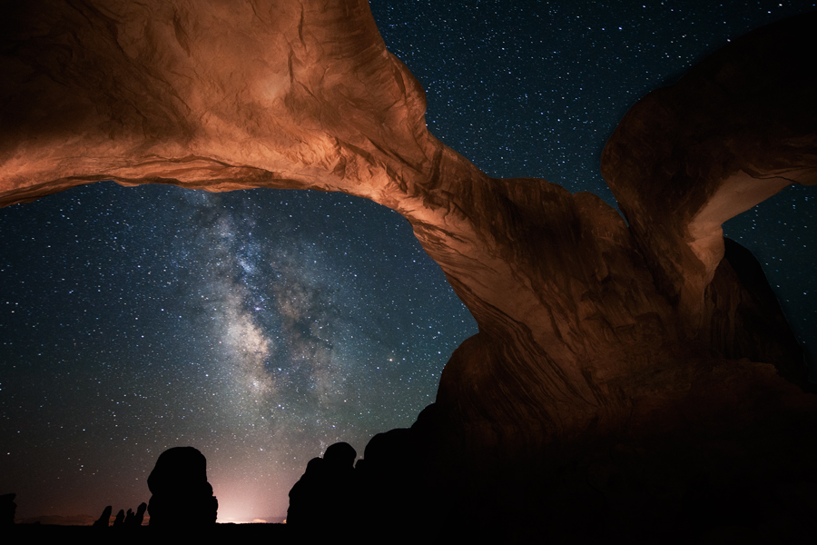 Unter einem schwach beleuchteten Steinbogen steigt die Milchstraße auf, dahinter zeichnen sich am Horizont die Silhouette von Felsen vor einem hellen Licht ab.