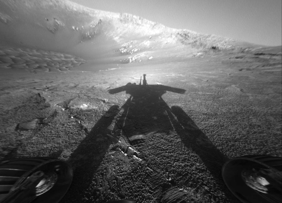 Der Schatten eines Marsrovers fällt auf steiniges Gelände, im Hintergrund ragt eine bergflanke auf.