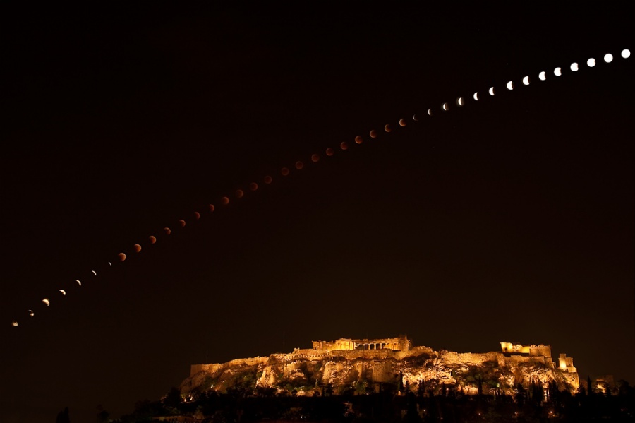 Über einem nächtlichen, beleuchteten Hügel mit historischen Gebäuden - der Akropolis - findet eine Mondfinsternis statt, die in vielen einzelnen Aufnahmen in einem Bogen über den schwarzen Himmel zieht.