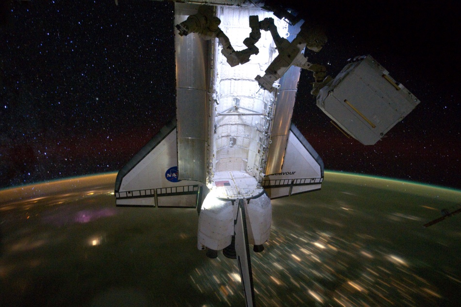 Die Raumfähre Endeavour schwebt aufgerichtet mit geöffneter Ladebucht, die innen beleuchtet ist, über der Erde, die unten vorbeizieht. Auf der Erde sind Stadtlichter als Streifen zu sehen.