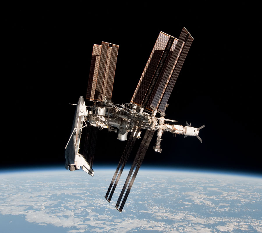 Die Raumfähre Endeavour ist mit offener Ladebucht an die Internationale Raumstation ISS angedockt, darunter ist die Erde mit Wolken und Meer.