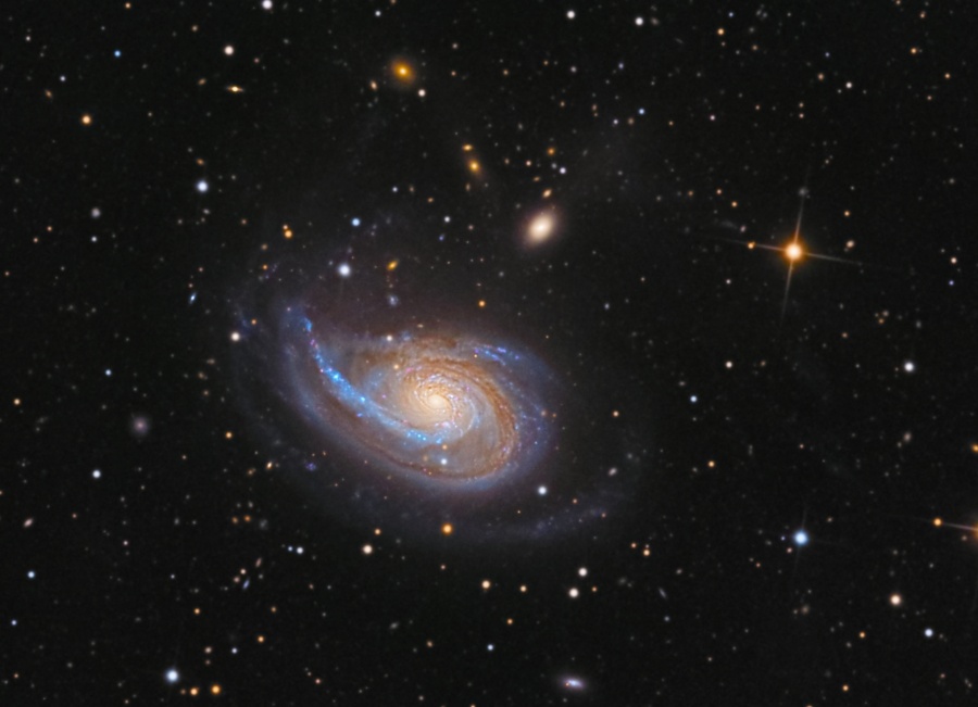 Die Galaxie in der Mitte hat eine Spiralform, doch nach links ist ein sehr mächtiger, markanter Spiralarm über die Galaxie hinausgezogen, der von blauen Sternhaufen gesäumt ist. Im Hintergrund sind Sterne und einige kleine Galaxien verteilt.