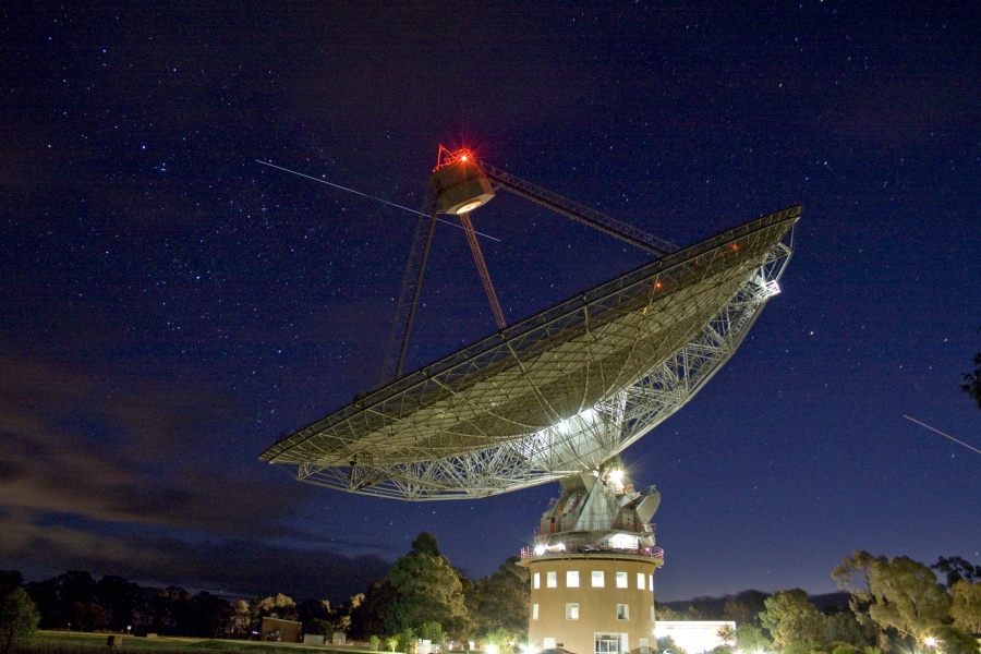 Im Vordergrund steht eine riesige Radioantenne, die von der Seite zu sehen und von unten beleuchtet ist. Am Fangspiegel der Antenne leuchtet ein rotes Licht. der Himmel dahinter ist dunkelblau mit Strichspuren.