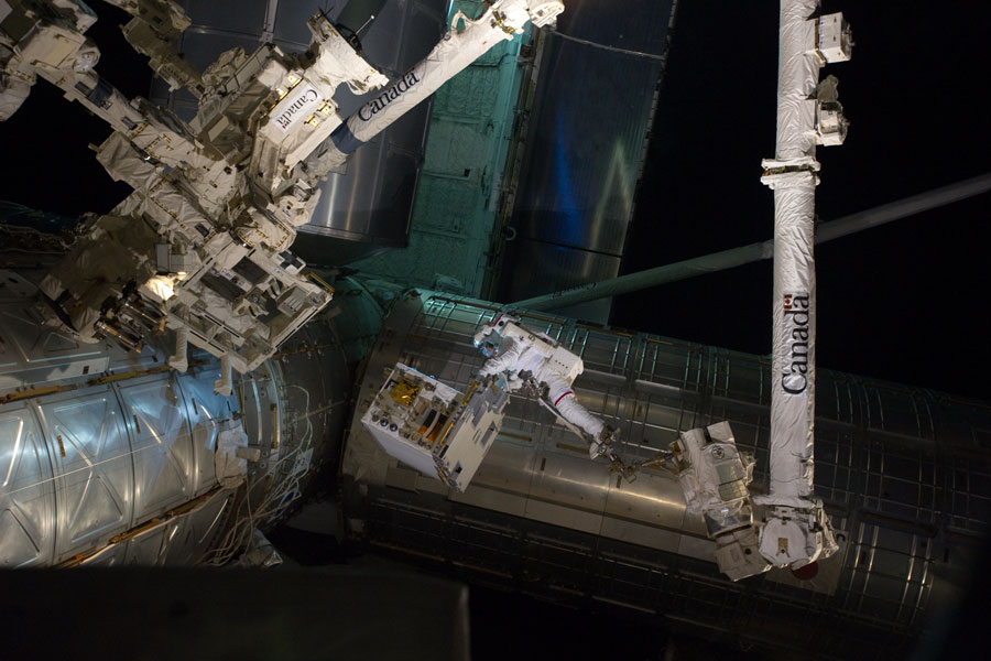 Das Bild zeigt Teile der Raumstation, einen Astronauten und den Canadarm bei Nacht.