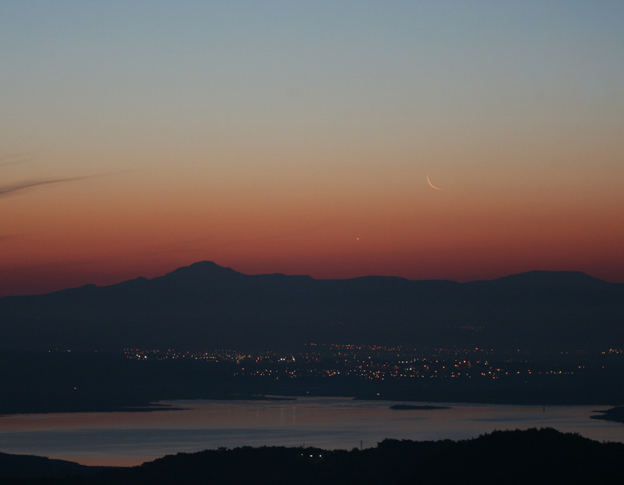 Im Vordergrund ist ein Stausee, dahinter eine Stadt und noch weiter entfernt ein Gebirge. Darüber leuchtet Abendrot, das in nellblau übergeht. An der Grenze des Abendrots steht eine schmale Mondsichel und der Planet Venus.