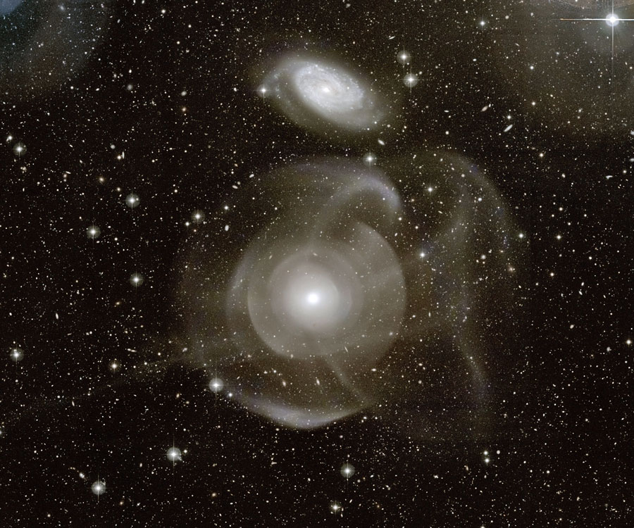 Zwei Galaxien im Bild wirken sehr gleichförmig, die obere ist kleiner, spiralförmiger und heller, die untere ist von eigenartigen Schalen umgeben.