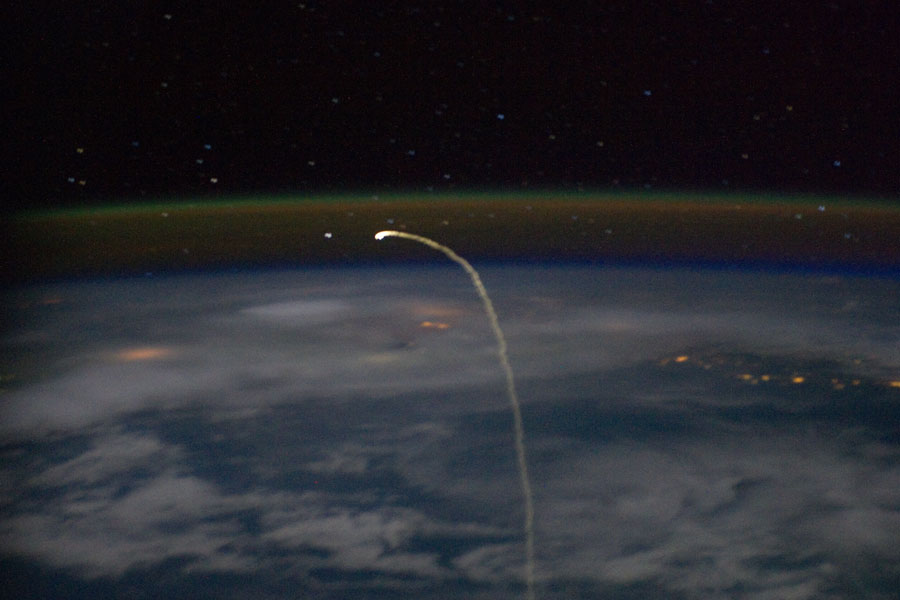 Über der Erde verläuft eine helle Spur nach oben und bildet einen Haken nach links. Über dem Erdrand leuchtet eine grünliche Schicht aus Nachthimmellicht.