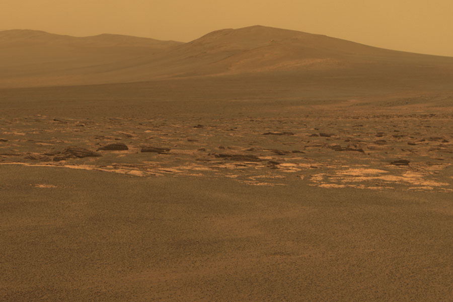 Eine vorne glatte rötlichbraune Marslandschaft geht in felsiges Gelände über, im Hintergrund ragen Berge vor einem rötlichen Himmel auf.