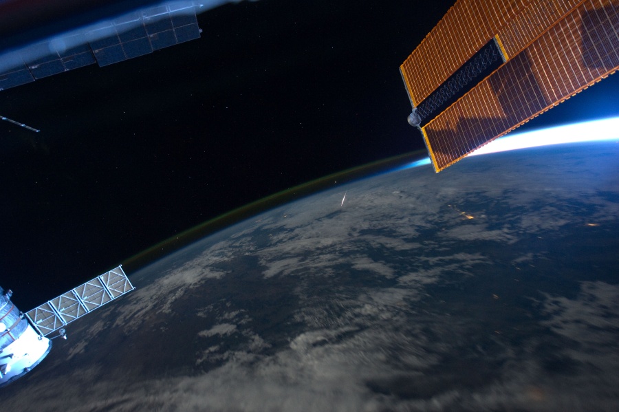 Über der dunklen Erde zeichnet sich ein zarter Streifen Nachthimmellicht ab. Rechts ist ein kurzes Stück der sonnenbeleuchteten Erdsichel, darüber ein Solarpaneel. Links ein Teil einer Sojus-Kapsel.