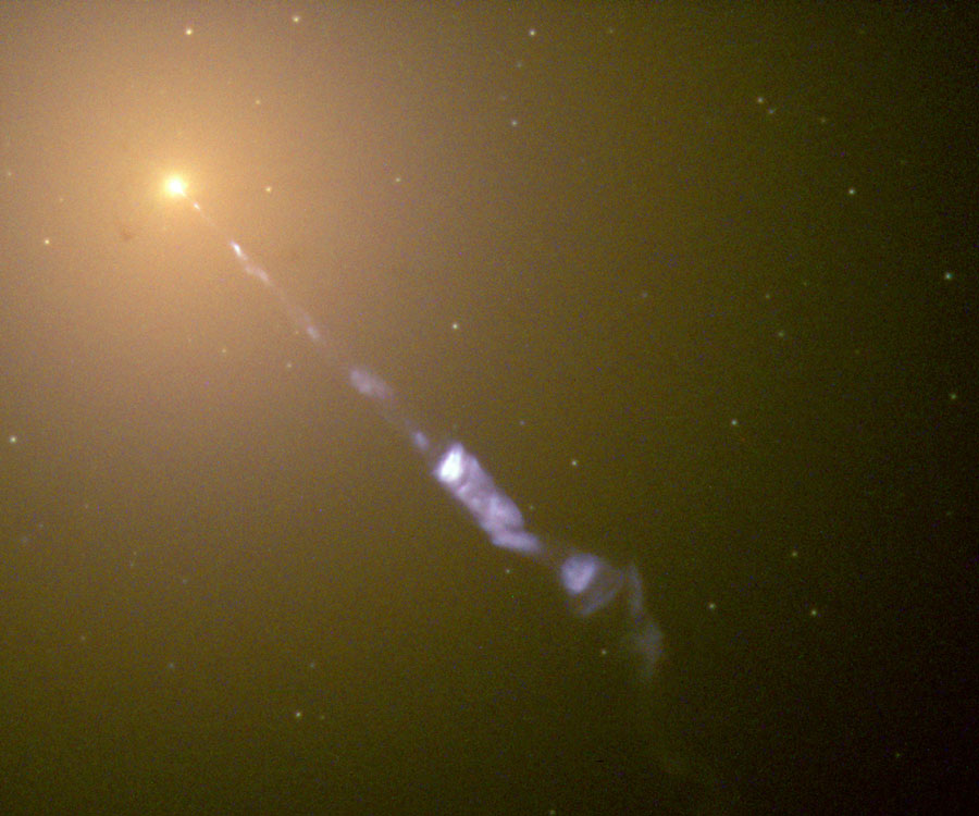 Links oben ist ein gelb leuchtendes Galaxienzentrum, von diesem strömt ein langer, blauer Strahl, der teilweise unterbrochen ist. 