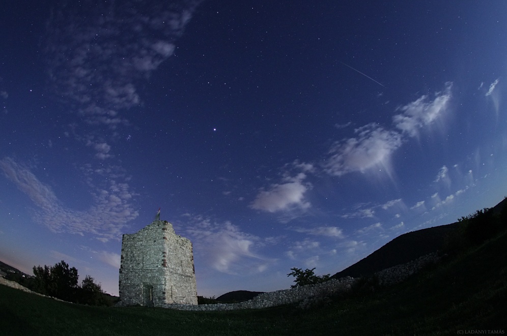 Hinter einem Turm im Mondlicht sind am nächtlichen Himmel einige Wolken, Sterne und Meteore zu sehen.