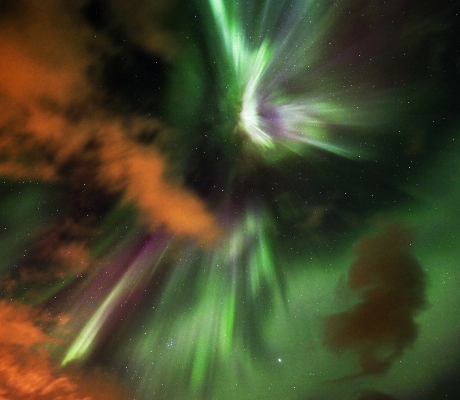 Das Bild zeigt sehr lebhafte grüne Polarlichter, die von der Mitte ausströmen, und rotbraune Wolken auf der linken Seite.
