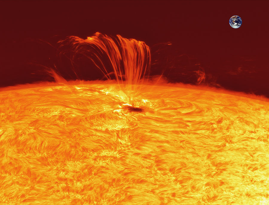 Aus einem kleinen Sonnenfleck strömen rote Plasmafäden entlang von Magnetlinien aus der Sonne. Der Sonnenfleck ist so groß wie die Erde, die im gleichen Maßstab rechts oben eingefügt ist.