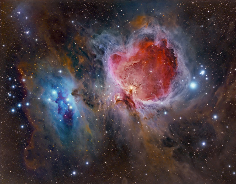 Mitten im Bild ist der Orionnebel in braune Staubwolken eingebettet und von markanten Sternen umgeben. Links ist ein blauer Reflexionsnebel, der Orionnebel selbst wirkt wie eine innen rot leuchtende Höhle.