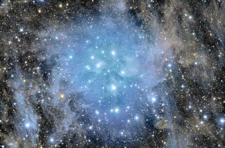 Mitten im Bild strahlt ein Sternhaufen, umgeben von leuchtend blauen Staubnebeln. In unmittelbarer Umgebung leuchten die Staubnebel blau, in größerer Entfernung sind die Nebel graubraun.