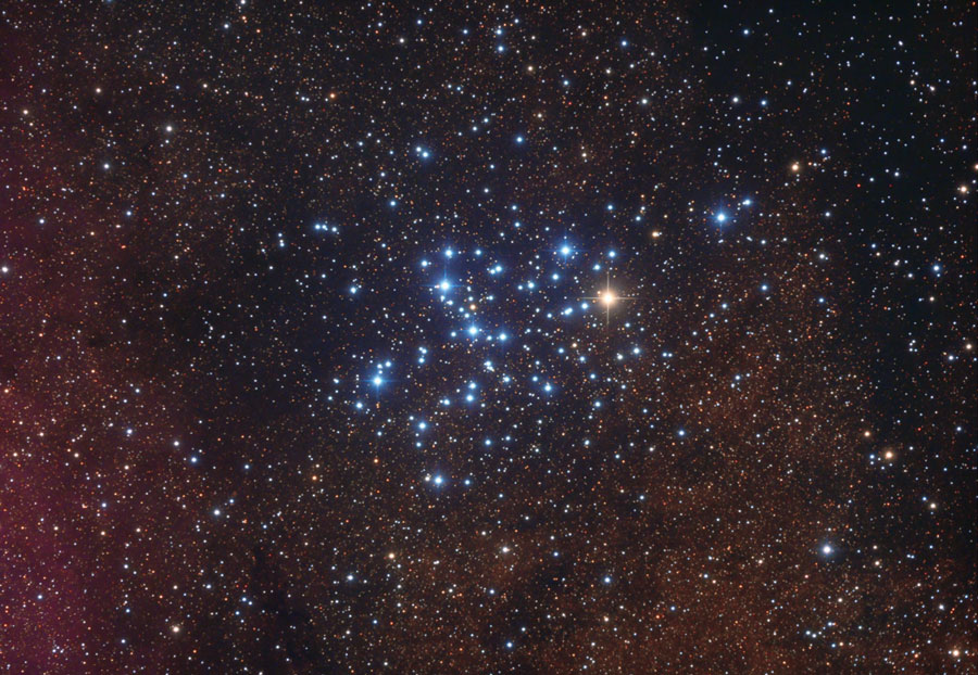 Die im Bild verteilten Sterne bilden in der Mitte einen Haufen aus helleren blauen Sternen. Ein Stern im Haufen leuchtet orangefarben.