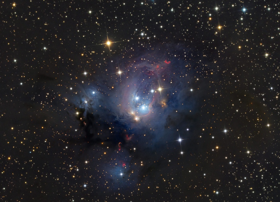 Im Bild zwischen glitzernden Sternen leuchtet ein blauer Nebel mit einigen roten Bögen. Die Bögen sind Herbig-Haro-Objekte, die aktive Sternbildung verraten.