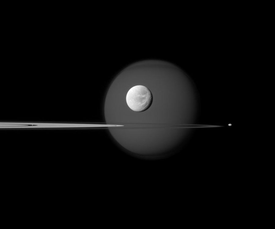Von links ragen die fast von der Seite sichtbaren Saturnringe ins Bild. Hinten ragt Titan groß auf, davor leuchtet Dione, rechts außerhalb der Ringe ist Pandora und in der Encketeilung ist der Schäfermond Pan.