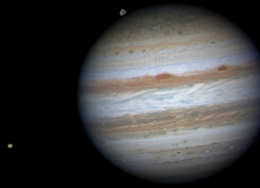 Der Planet Jupiter mit Wolkenbändern füllt rechts das Bild bis über den Rand hinaus, links ist der vulkanische Mond Io am Bildrand. Oben raucht Ganymed hinter Jupiter auf.