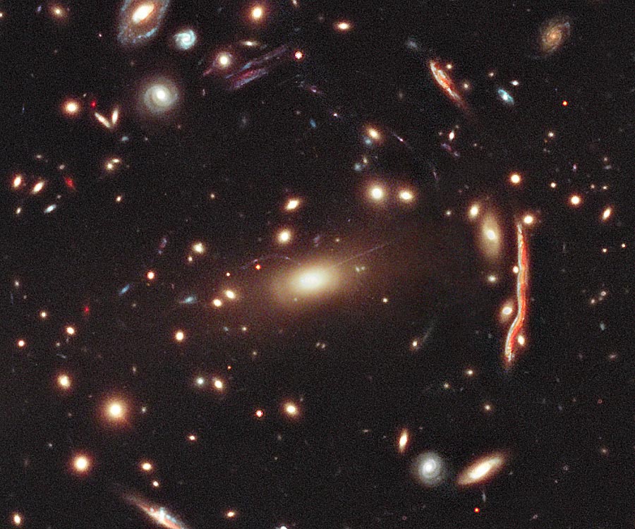 Das Bild ist voller Galaxien. In einem Kreis sind verzerrte Bilder von weiter entfernten Galaxien um eine helle Galaxie in der Mitte verteilt.