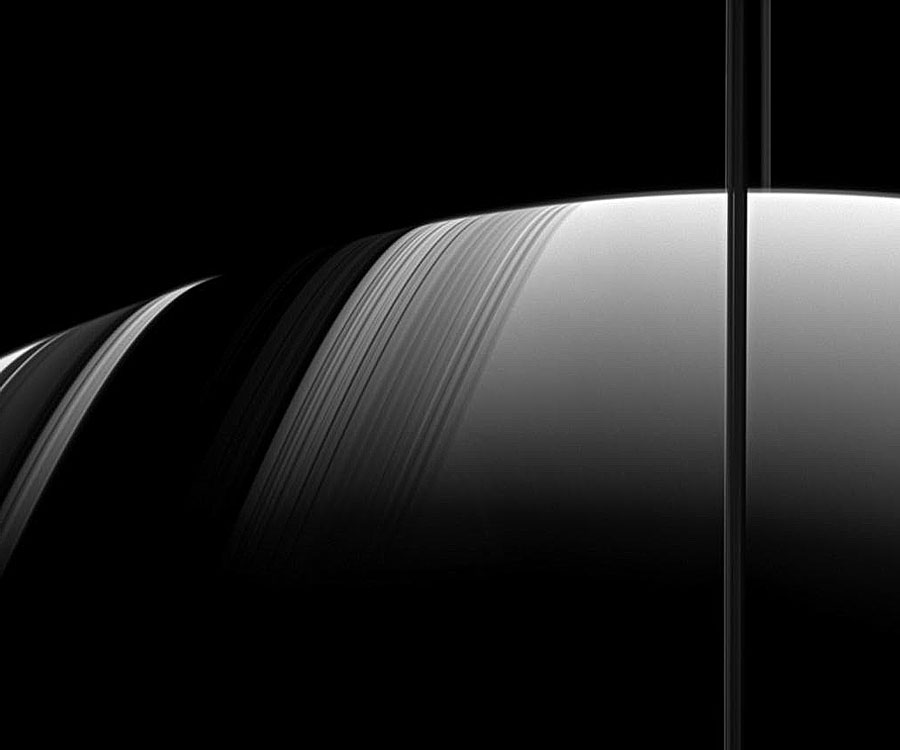 Der Ringplanet Saturn liegt scheinbar auf der Seite, oben ist der Rand, die Ringe laufen als schwarzer Streifen senkrecht rechts durchs Bild, links sind die Schatten auf dem Planeten zu sehen. Das Bild ist schwarzweiß.