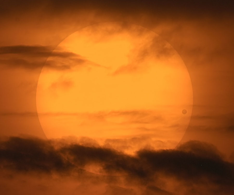 Hinter Wolken zeichnet sich die Sonne am orangefarbenen Himmel ab, rechts ist der Planet Venus als dunklere Scheibe zu sehen.