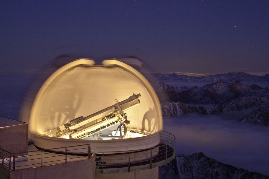 Die Teleskopkuppel im Bild ist scheinbar weit aufgeklappt und innen beleuchtet, im Inneren steht ein Teleskop. Der Trick ist, das Bild lang zu belichten und dabei die Kuppel zu drehen. Im Hintergrund ist eine verschneite Nachtlandschaft im Gebirge.