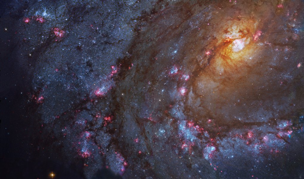 Links oben ist das gelbliche helle Zentrum einer Galaxie, ihre Spiralarme breiten sich nach links unten auf. Ein Spiralarm verläuft scheinbar über das Zentrum der Galaxie. In den Spiralarmen sind viele blaue Sternhaufen und rosarote Sternbildungsgebiete.