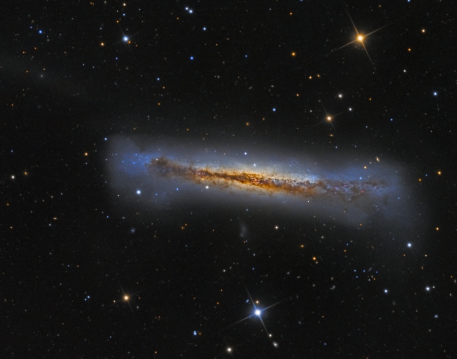 Die Galaxie im Bild ist von der Kante sichtbar, sie wirkt fluffig und aufgebauscht. In der Mitte verläuft ein orangebrauner Staubwulst über eine gelblich leuchtende galaktische Ebene, außen ist sie von einem blauen Nebel umgeben.