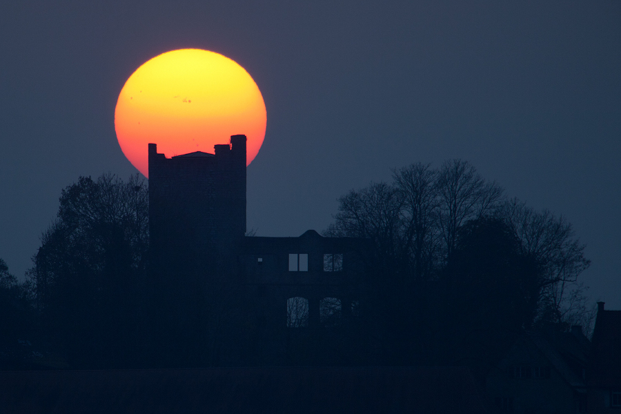 Hinter der Silhouette einer Ruine leuchtet eine gelborangefarbene Sonne in der Dämmerung.