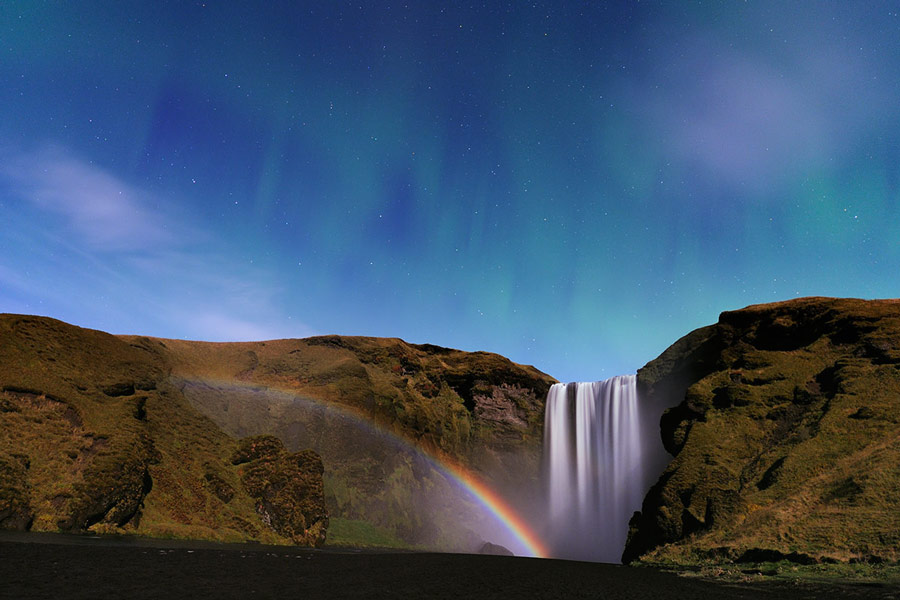 Über einen Steilhang stürzt ein Wasserfall. Nach links breitet sich ein Regenbogen aus.