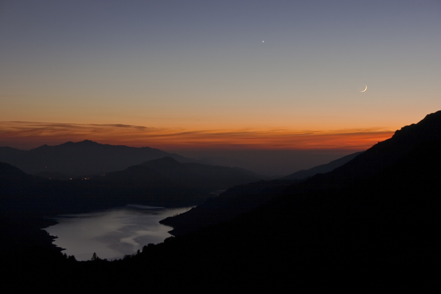 Im Abendrot in der Dämmerung leuchtet der Sichelmond im Westen. Etwas höher leuchtet die Venus. Links unten liegt ein ruhiger See am Fuß eines Berghanges, der rechts im Bild als Silhouette erkennbar ist.