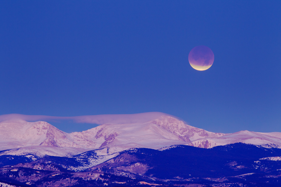 Über einem verschneiten Gebirge leuchtet am blauen dunklen Himmel in der Dämmerung der Mond. Er ist oben rot und verfinstert, die untere Kuppe ist von der Sonne beleuchtet.