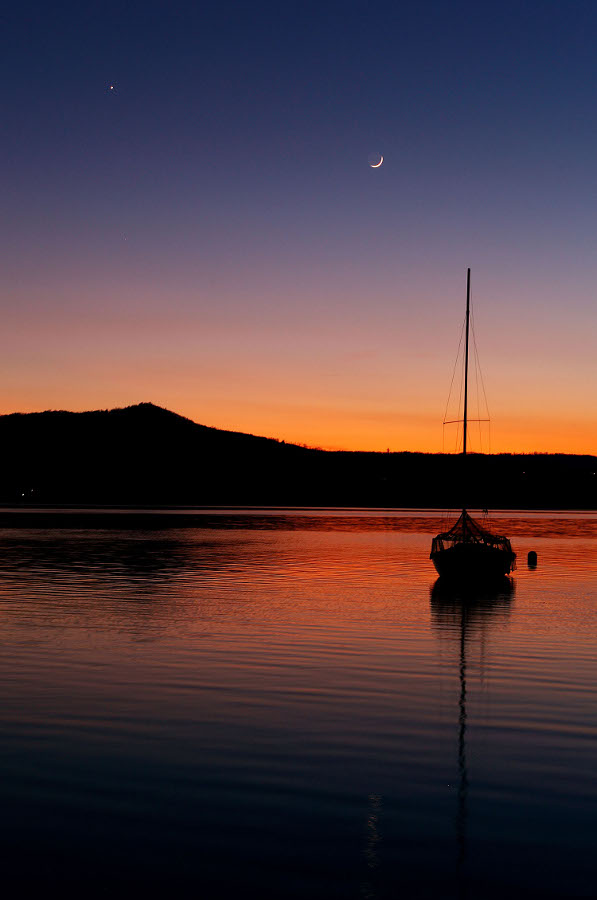 Auf einem ruhigen See, der in der Abenddämmerung rot leuchtet, schwimmt ein Schiff. Hinten ist die Silhouette eines Berges. Am violettblauen Himmel leuchtet ein junger Sichelmond.