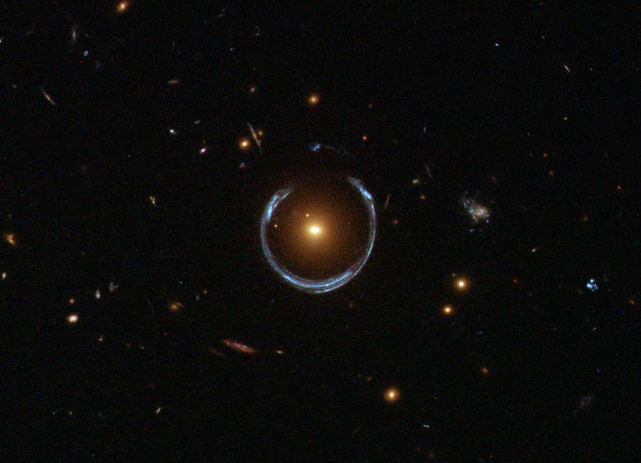Um eine elliptische Galaxie verlaufen blaue Bögen. Im Hintergrund sind weitere kleine Galaxien verteilt.