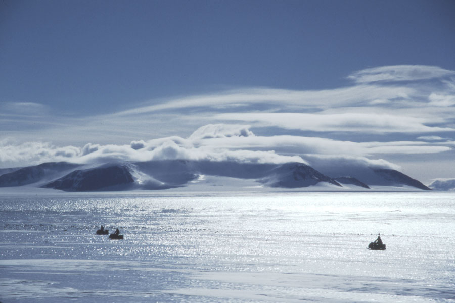Die Landschaft ist eisbedeckt und glitzert in der Sonne. Drei Silhouetten von Schneefahrzeugen sind zu sehen. Im Hintergrund ragen Berge auf, darüber ziehen einige Wolken über den blauen Himmel.