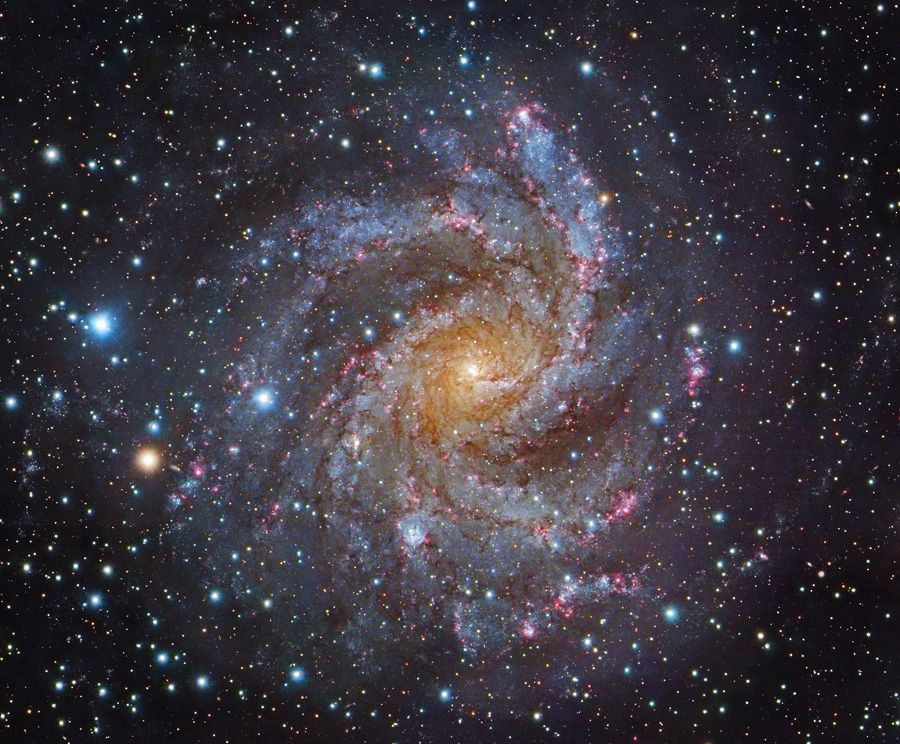 Die Galaxie im Bild ist direkt von oben zu sehen. Sie hat einen gelben Kern und ist von flockigen, blauen Spiralarmen umgeben, in denen rosarote Sternbildungsgebiete verteilt sind. In ihrer Umgebung sind viele Sterne zu sehen.