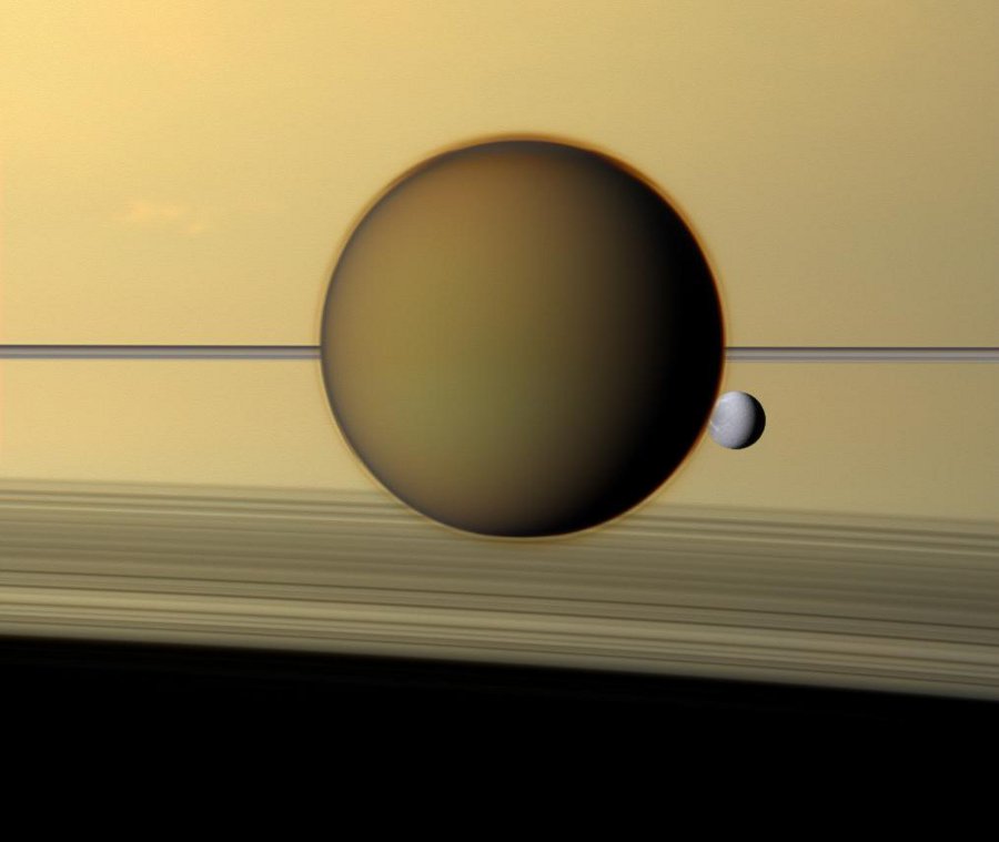 Vor dem Planeten Saturn, der das Bild ockerfarben füllt, ziehen die Ringe einen Strich durchs Bild und werfen nach unten einen Schatten auf den Planeten. Davor ist der riesige Mond Titan und dahinter der kleinere Mond Dione.