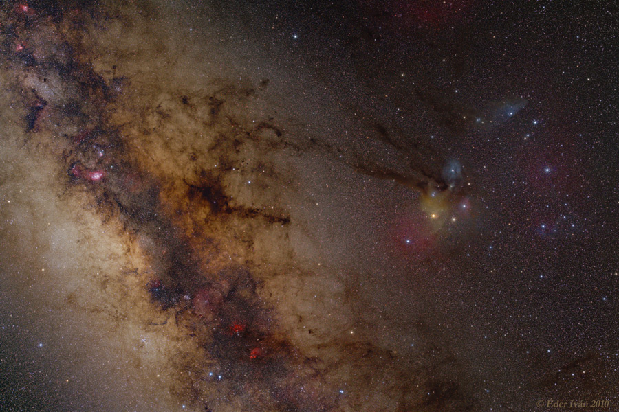 Links im Bild zieht eine dunkle Staubwolke über das nebelige Leuchten der Milchstraße. Rechts daneben sind einige Sterne, die von bunten Nebeln umgeben sind.