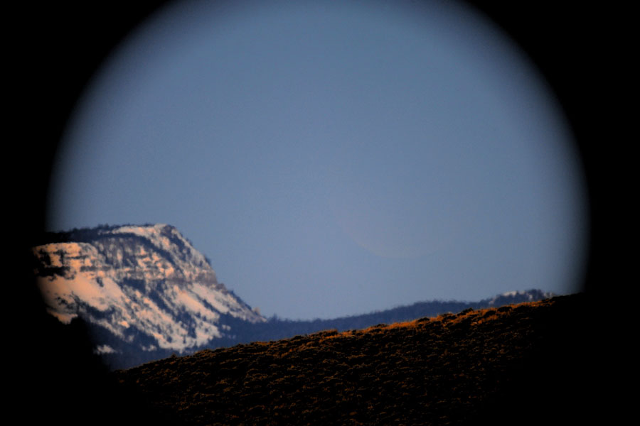 Mitten im Bild steht der Mond, aber er ist kaum erkennbar. Außen ist eine dunkle Blende, links ein Berg. Der Himmel ist blau von der Dämmerung.