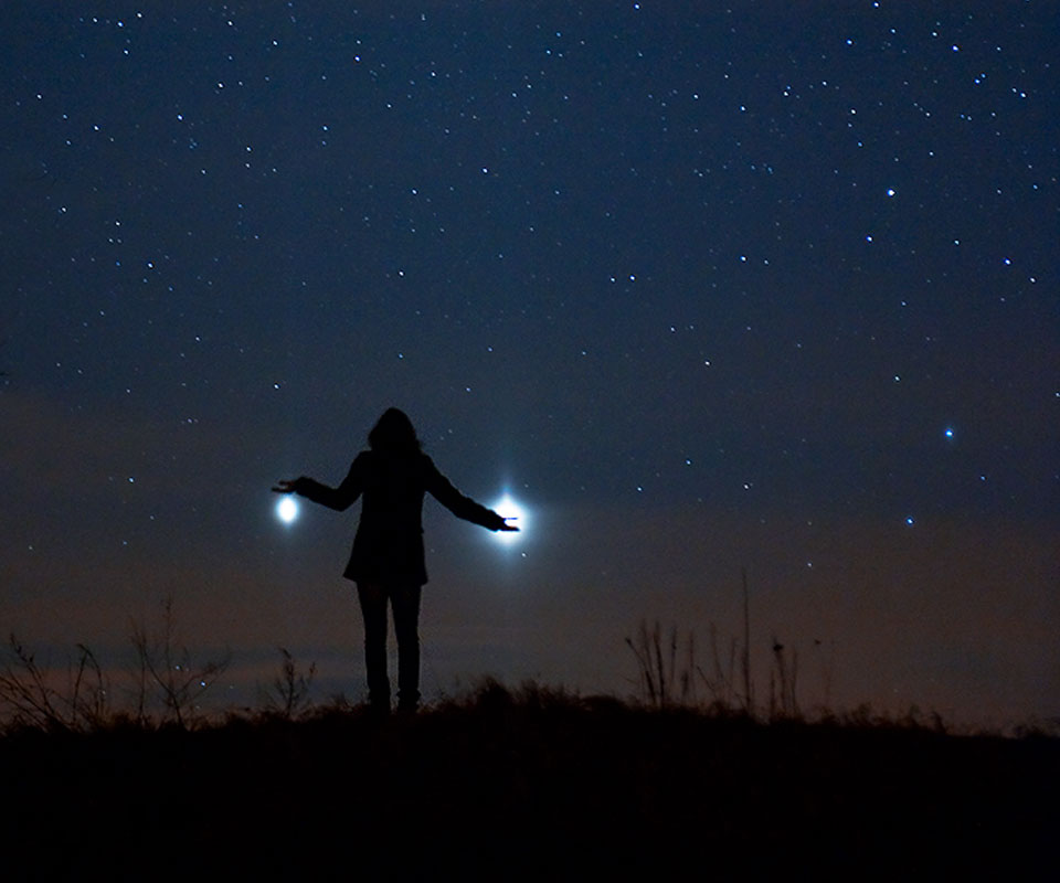 Vor einem sternklaren Himmel zeichnet sich die Silhouette einer Person ab. Sie hält scheinbar links und rechts helle Lichter in der Hand, es sind die Planeten Jupiter und Venus.