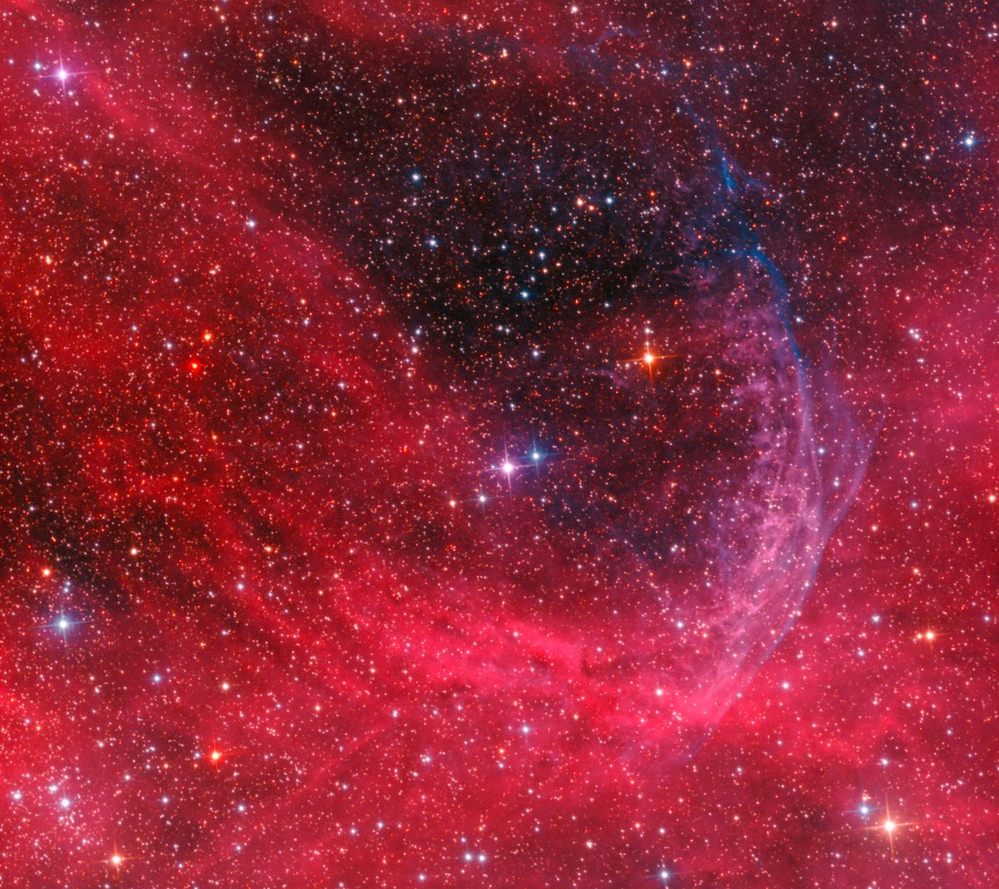 In einem rot leuchtenden Nebelfeld mit vielen Sternen zeichnet sich eine halbringförmige Struktur ab.