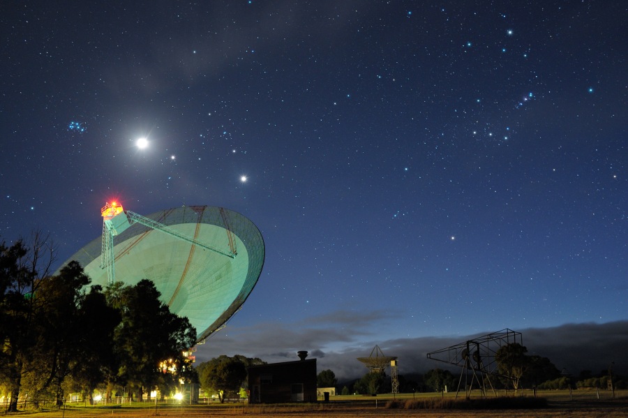 Hinter Bäumen ragt eine grün beleuchtete Radioantenne auf, dahinter sind die Sternbilder Stier und Orion markiert. Mariante Objekte sind die Plejaden, Mond, Jupiter und Venus.