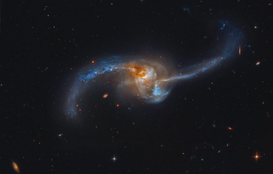 Im Zentrum ist ein wirres Knäul, das aus zwei Galaxien besteht, links ist ein gekrümmter Schweif aus Sternen, Staub und blauen Sternhaufen hinausgeschleudert, rechts ein relativ gerader Arm aus Sternen und Staub.