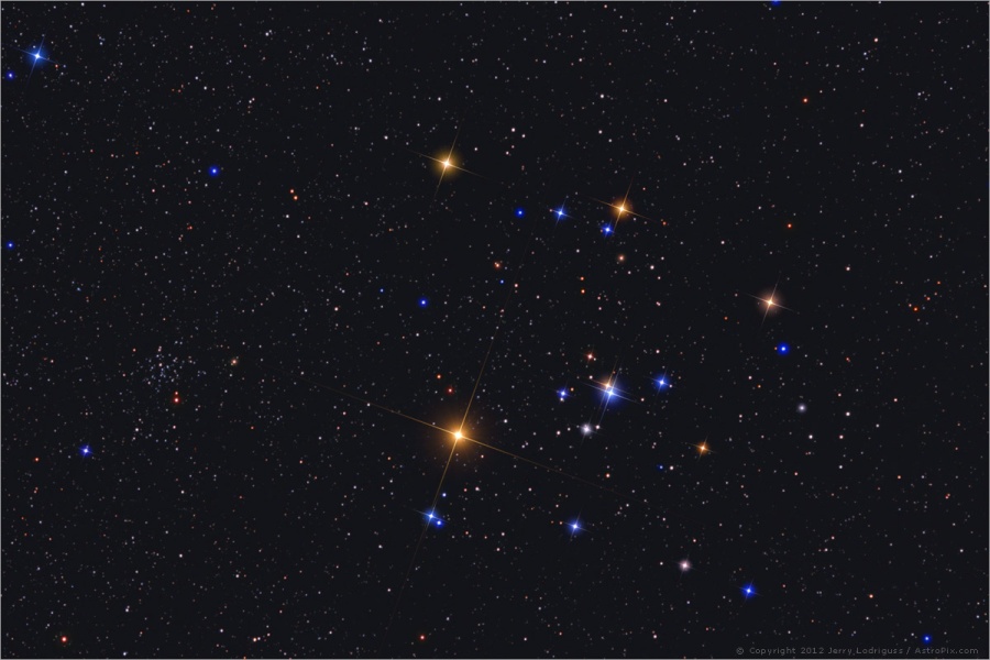 Die Hyaden sind ein kompakter, großer Sternhaufen in der Bildmitte. Links leuchten zwei rote, hellere Sterne, rechts daneben sind viele Sterne V-förmig verteilt.